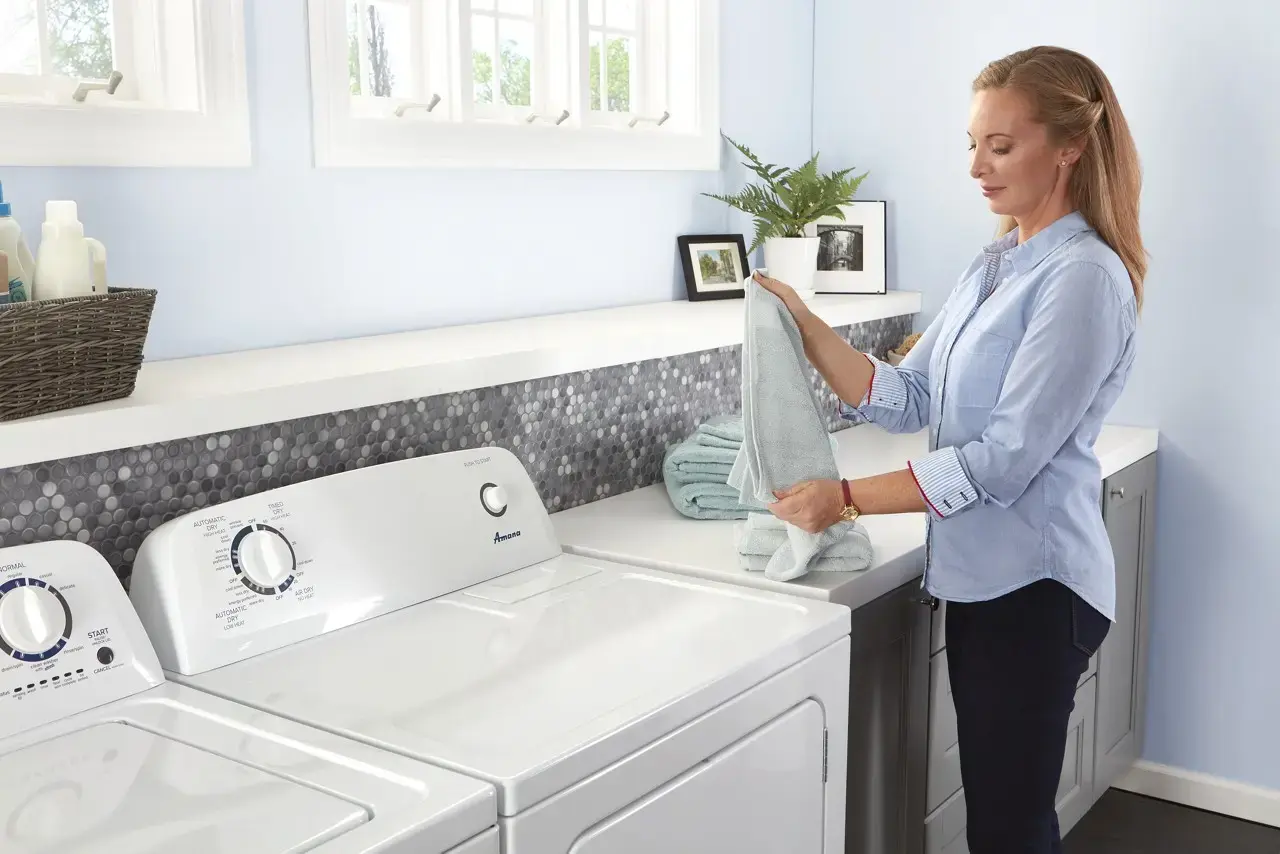 Woman folding a light gray towel beside a white washing machine and dryer in a bright laundry room.