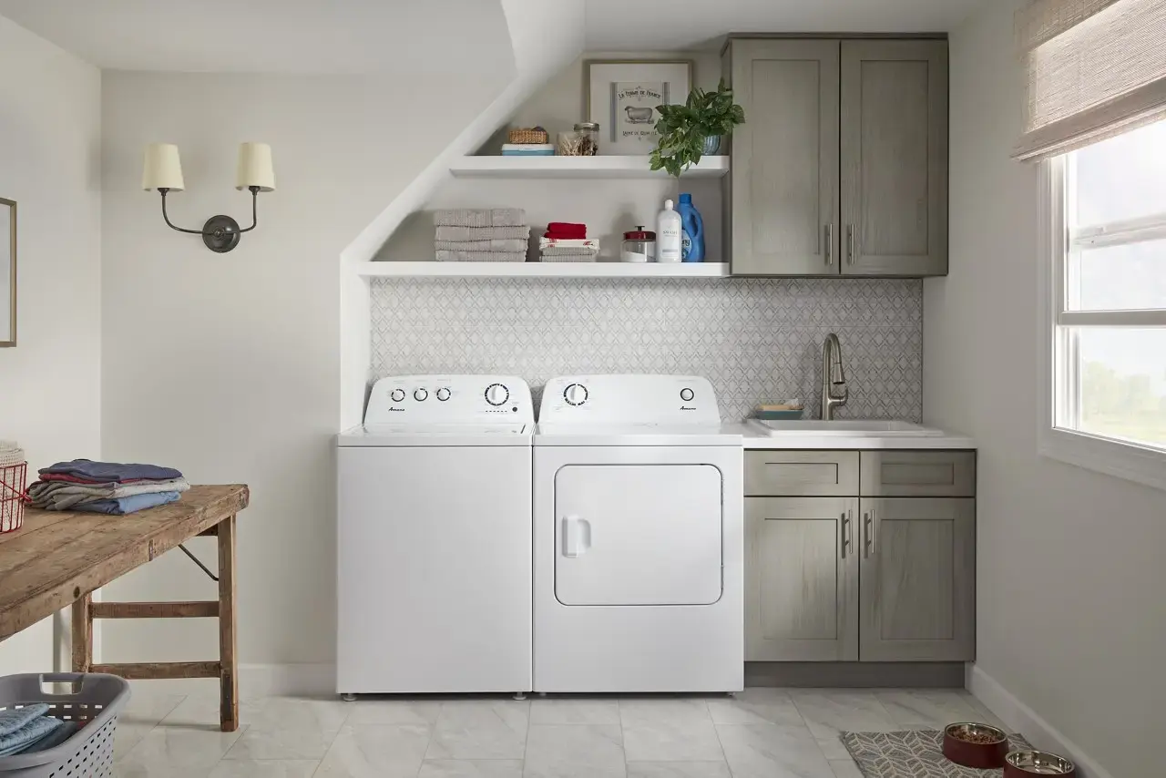 White washer and dryer side by side under a shelf, with gray kitchen-style cabinets and a utility sink on the right in a bright laundry room.
