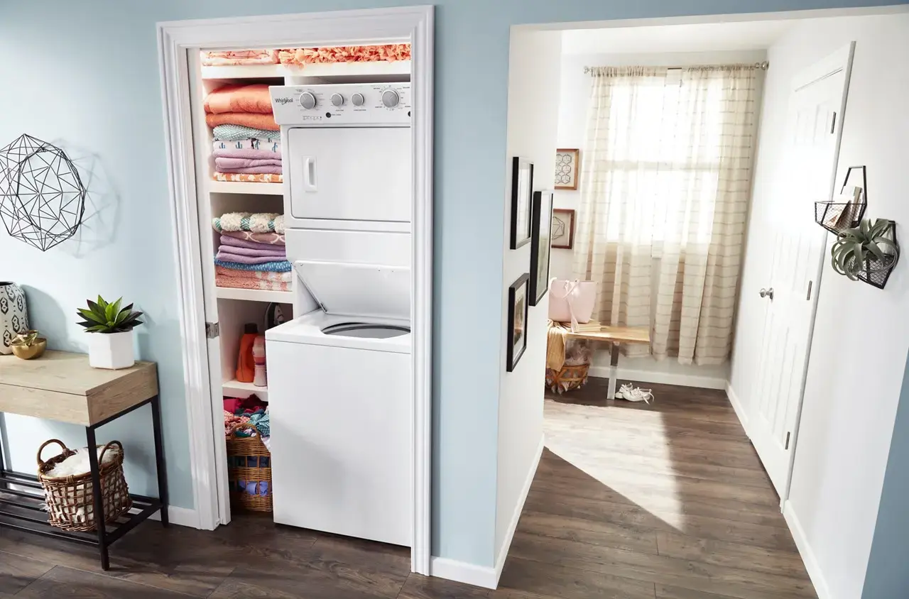 Closet with a stacked washer and dryer and shelves of folded towels; doorway opens to a bright hallway.
