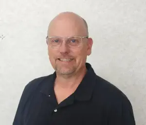 Portrait of a smiling bald man with glasses wearing a black polo shirt, posed against a light gray backdrop. (head-and-shoulders)