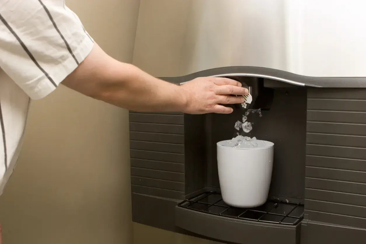 Hand placing ice into a white cup from a built-in ice dispenser in a dark coffee machine.
