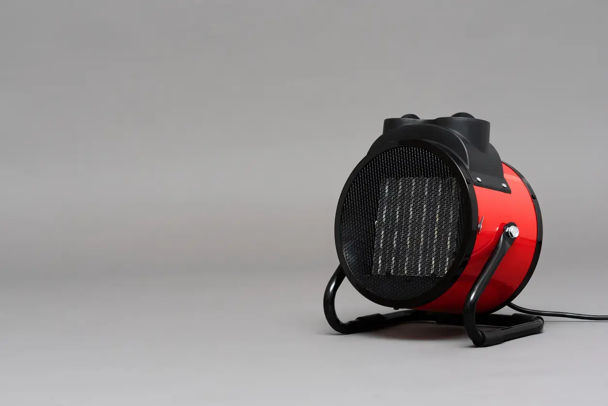 Red portable space heater with a black protective grille and two curved legs on a gray studio backdrop, cord to the side.